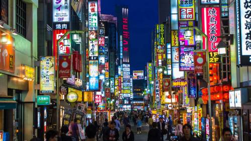 The bustling nightlife of Roppongi, Tokyo, with crowds of people on the street and buildings covered in vibrant neon signs.